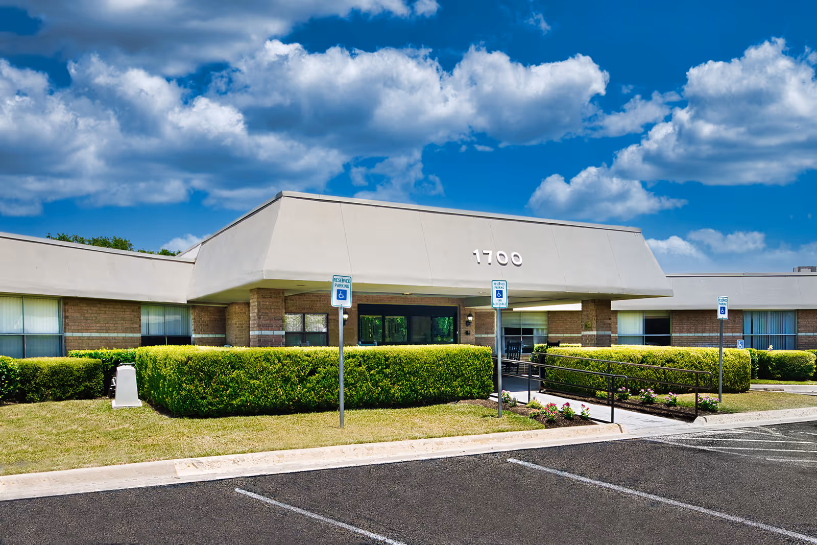 Front entrance of a single-story nursing facility with a canopy marked "1700", handicap parking signs, a wheelchair ramp, and trimmed hedges under a blue sky.