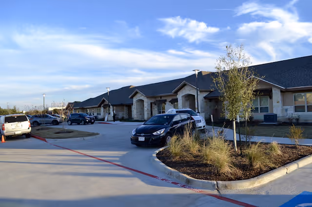 Exterior view of a single-story senior living facility building with a row of entrances, a parking lot with several parked cars, landscaped areas with small trees and shrubs, under a partly cloudy blue sky.