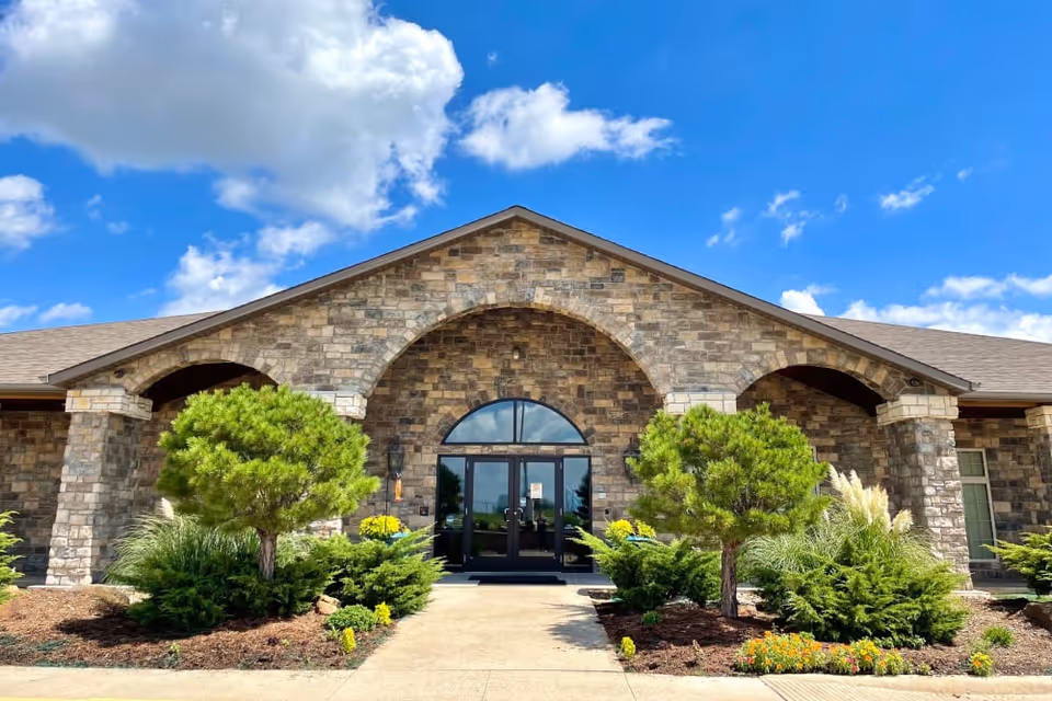 Front exterior view of a stone building with an arched entrance and double glass doors. The building is surrounded by landscaped greenery including small trees, bushes, and flowers under a bright blue sky with scattered clouds.