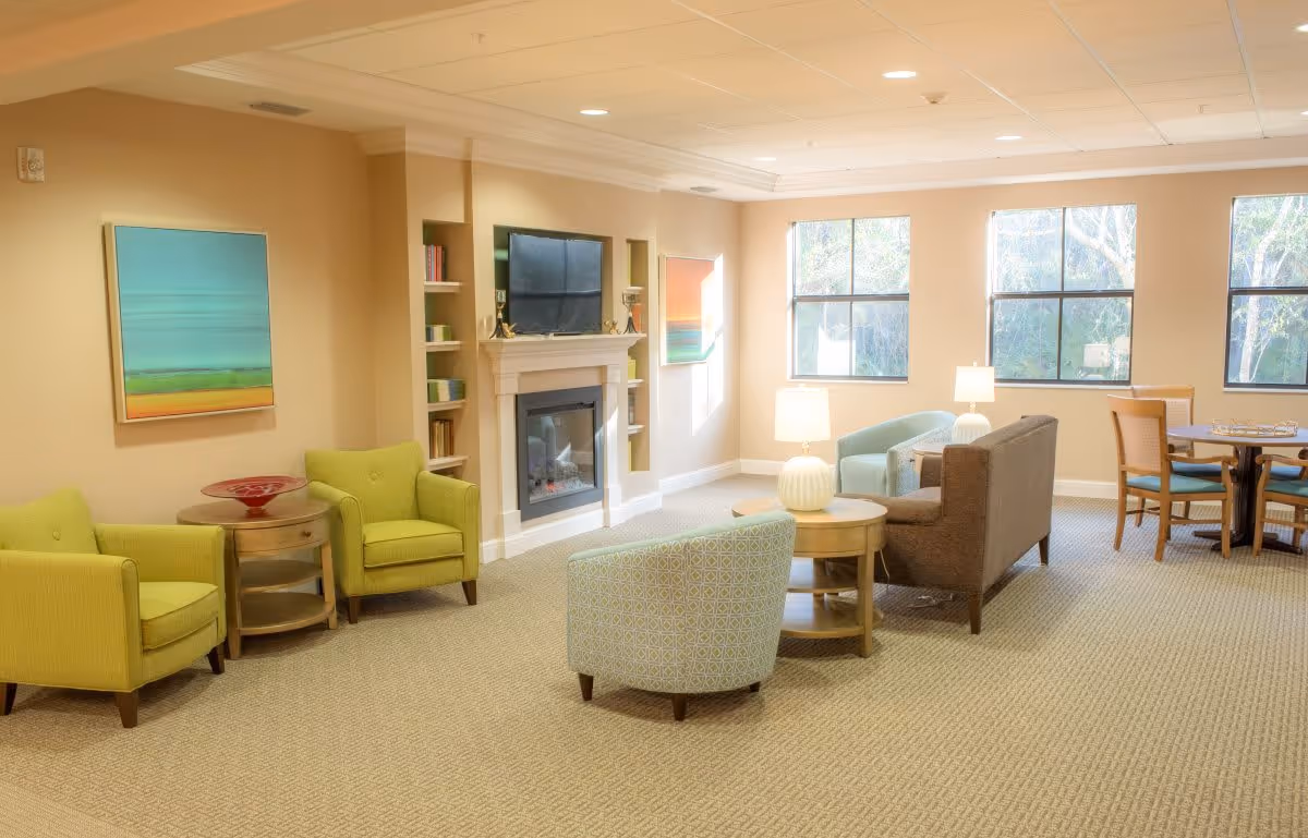 A bright and spacious living room area with beige walls and carpeted floor. The room features two green armchairs with a round wooden side table between them, a fireplace with a TV mounted above it, built-in shelves with books and decor, a brown sofa, a light blue armchair, two round wooden coffee tables with white lamps, and a dining table with four chairs near three large windows letting in natural light. Colorful abstract paintings hang on the walls.