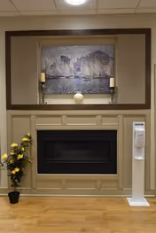 Interior view of a room featuring a beige fireplace with a black insert, a painting of a harbor scene above it, two candle holders with candles on the mantel, a potted plant with yellow flowers to the left, and a white hand sanitizer dispenser to the right.