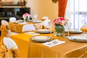 Dining room tables set with gold tablecloths, place settings, and floral centerpieces near windows with red curtains.