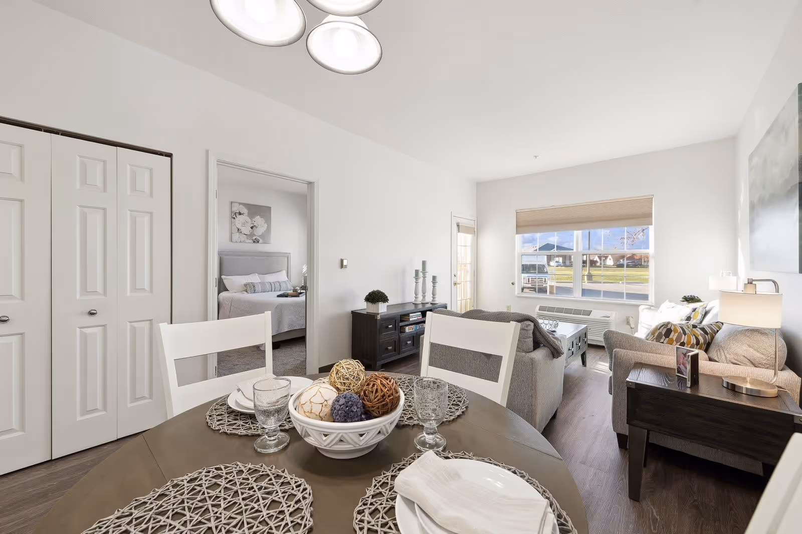 A bright and modern senior living apartment interior featuring a dining table set with plates, glasses, and decorative centerpiece in the foreground. Beyond the dining area is a cozy living room with a sofa, armchair, coffee table, and side table with a lamp. A large window lets in natural light and offers a view of the outside. A doorway leads to a bedroom with a neatly made bed and floral artwork above the headboard. The walls are white, and the flooring is wood-style.