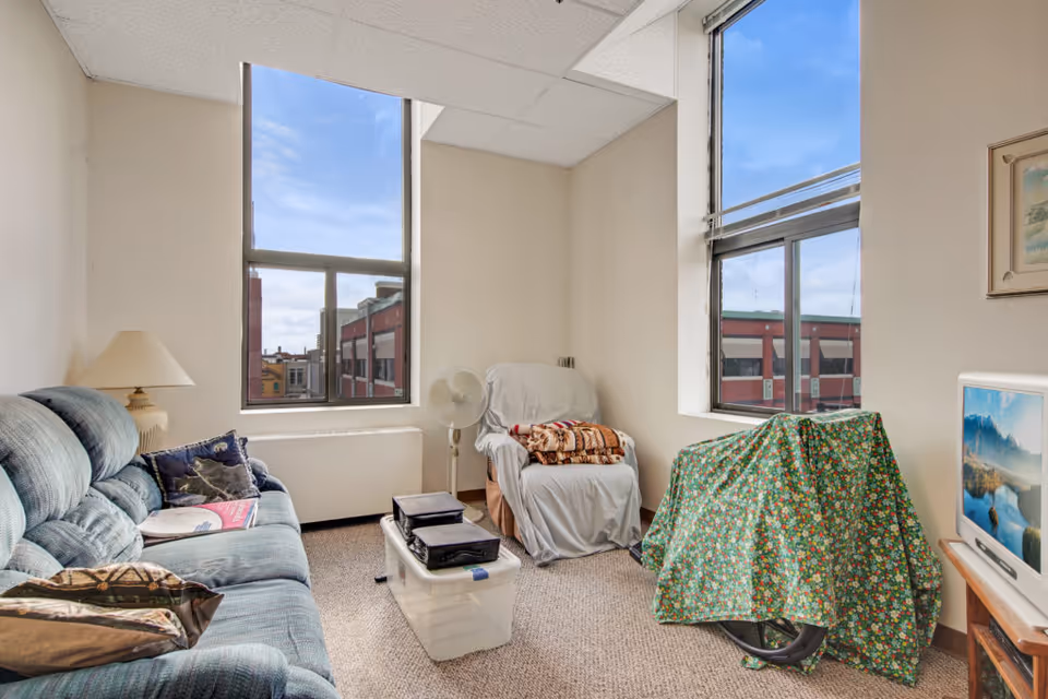 A small living room with a blue upholstered couch on the left, a beige armchair covered with a white sheet and a folded blanket on top, a standing fan, and a plastic storage bin with items on it. There is a bicycle covered with a green floral cloth near a window on the right. Two large windows show a view of nearby buildings under a blue sky with some clouds. A small TV on a wooden stand displays a scenic mountain and lake image.