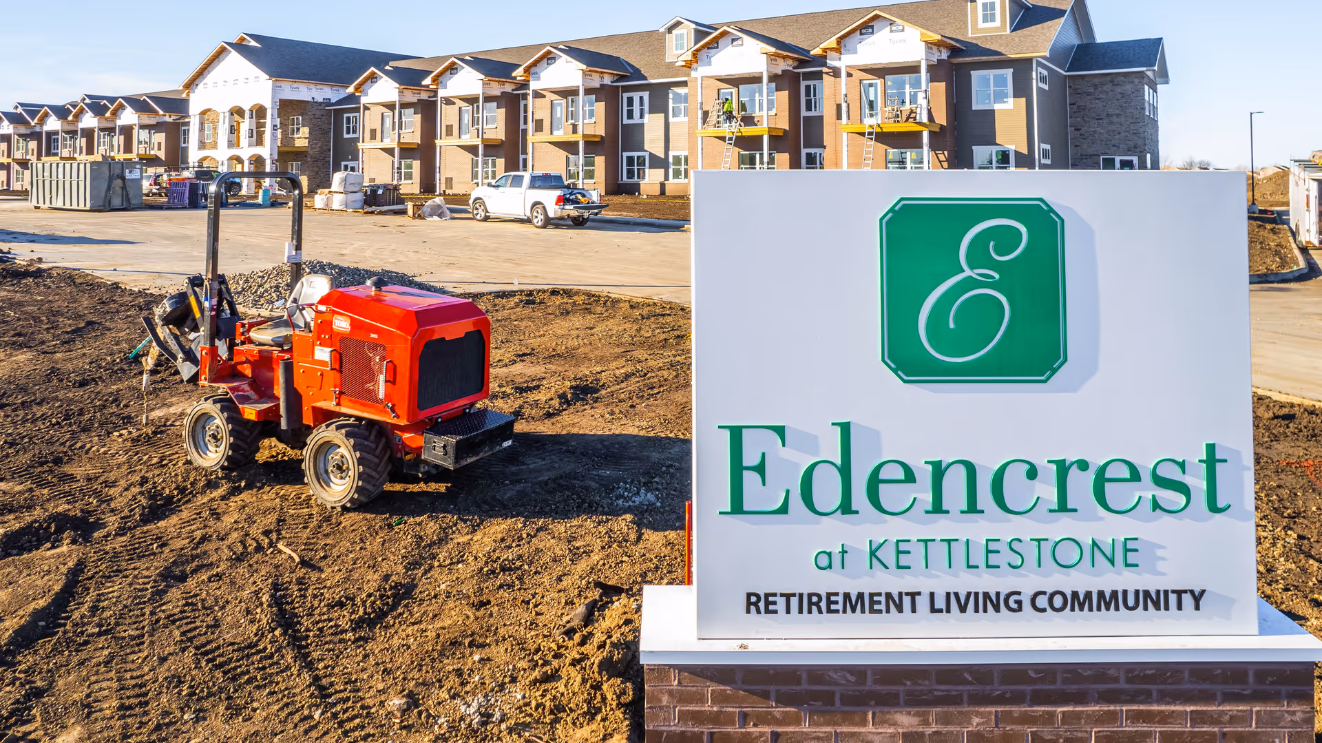 A large sign reading "Edencrest at Kettlestone Retirement Living Community" stands in front of a newly constructed apartment building with a small red construction vehicle on the dirt lot.