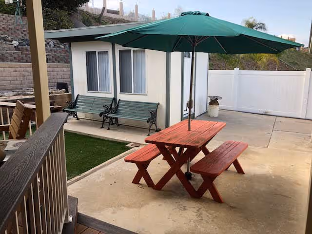 Outdoor patio area with a red wooden picnic table and benches under a large green umbrella. Two green benches are placed against a small white building with windows. The area is surrounded by a concrete floor, a white fence, and a retaining wall with plants on a slope.
