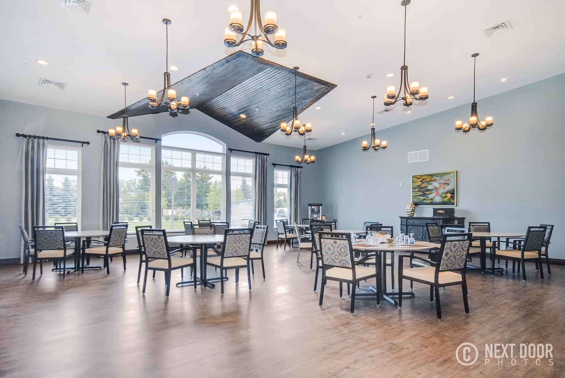 A spacious dining room with multiple round tables and chairs arranged neatly. The room features large windows with curtains allowing natural light to fill the space. Several chandeliers hang from the ceiling, and there is a decorative painting on the wall above a sideboard.