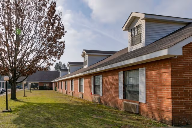 Exterior view of a single-story brick healthcare building with dormer windows, a grassy lawn, and a tree.