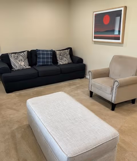 A cozy living room with a dark blue sofa adorned with three decorative pillows, a beige armchair with nailhead trim, and a light-colored rectangular ottoman in the center. A framed artwork featuring a red sun and dark landscape hangs on the beige wall behind the armchair.