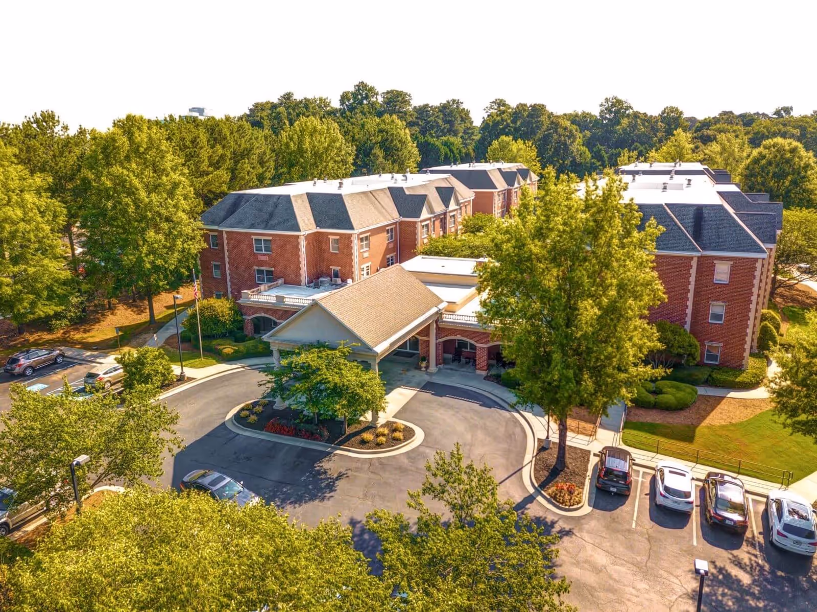 Aerial view of Dunwoody Place senior living facility showing a large brick building with multiple wings surrounded by trees and greenery. The entrance has a covered drop-off area with a circular driveway and several parked cars nearby.