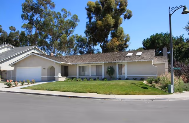 Single-story residential building with a sloped roof, garage, and front lawn with shrubs and trees under a clear blue sky.