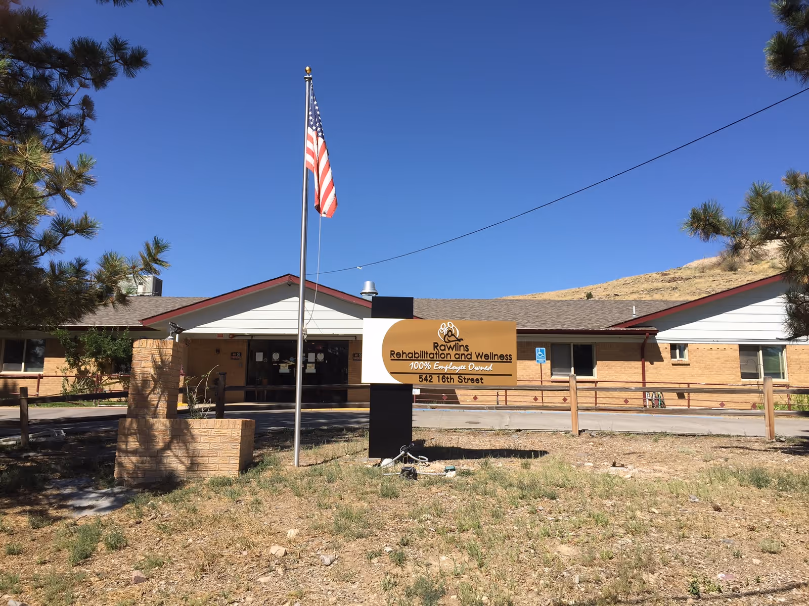 Exterior view of Rawlins Rehabilitation and Wellness building with a clear blue sky, an American flag on a flagpole, and a sign displaying the facility's name and address in front of the building. The building is single-story with a brick facade and a sloped roof, surrounded by some trees and dry grass.