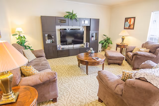 A cozy living room with beige patterned carpet, brown upholstered sofas and armchairs arranged around a round wooden coffee table. A large dark wood entertainment center with a flat-screen TV and decorative plants is against the wall. Two table lamps provide warm lighting, and a framed artwork hangs on the wall near a window with blinds.