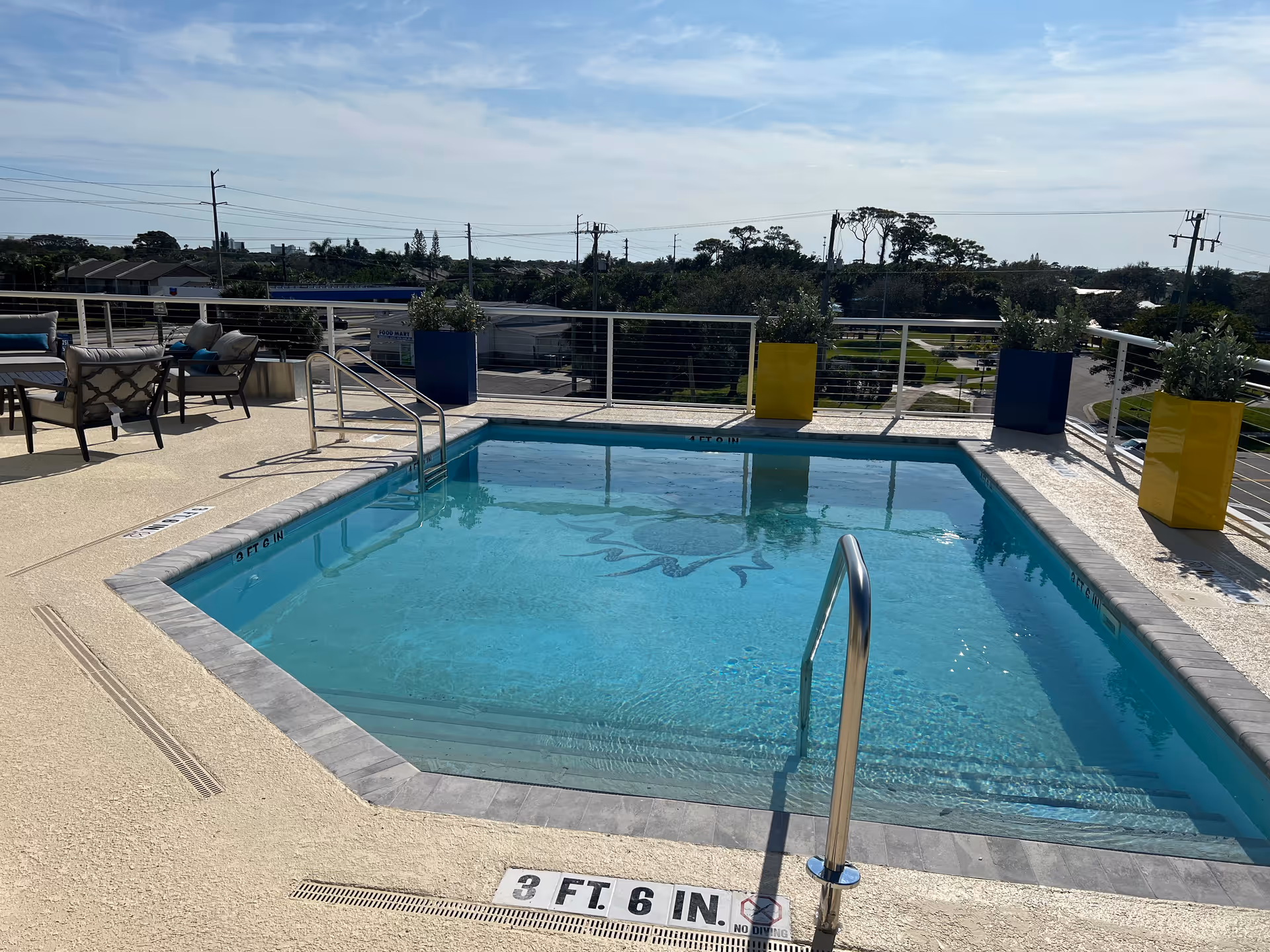 Outdoor swimming pool on a rooftop with clear blue water, surrounded by a safety railing and several large planters with greenery. There are chairs and tables with cushions on the left side of the pool. The sky is partly cloudy and trees and buildings are visible in the background.