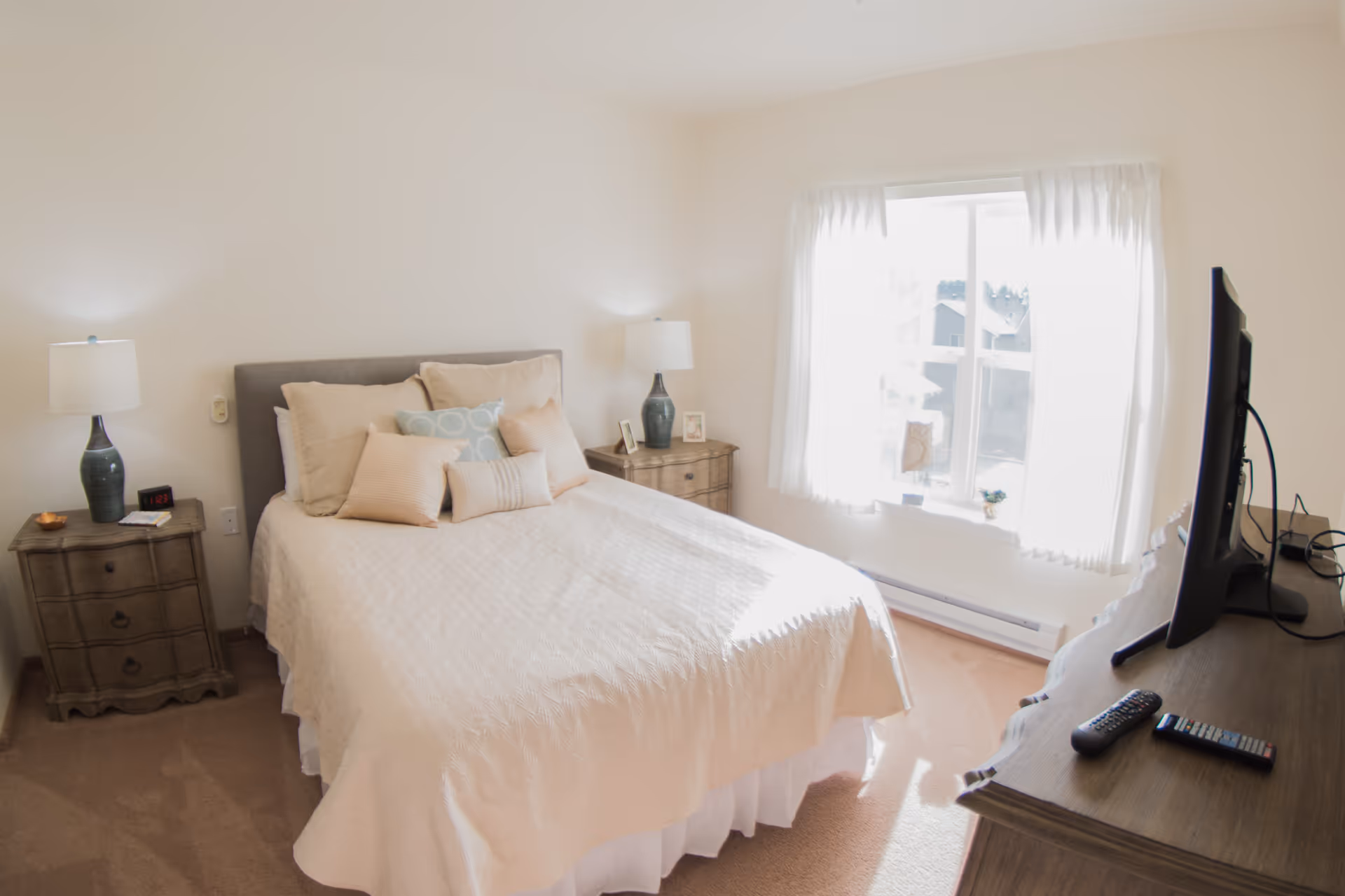 Bright furnished bedroom with a bed, matching nightstands and lamps, a window with curtains, and a TV on a dresser.