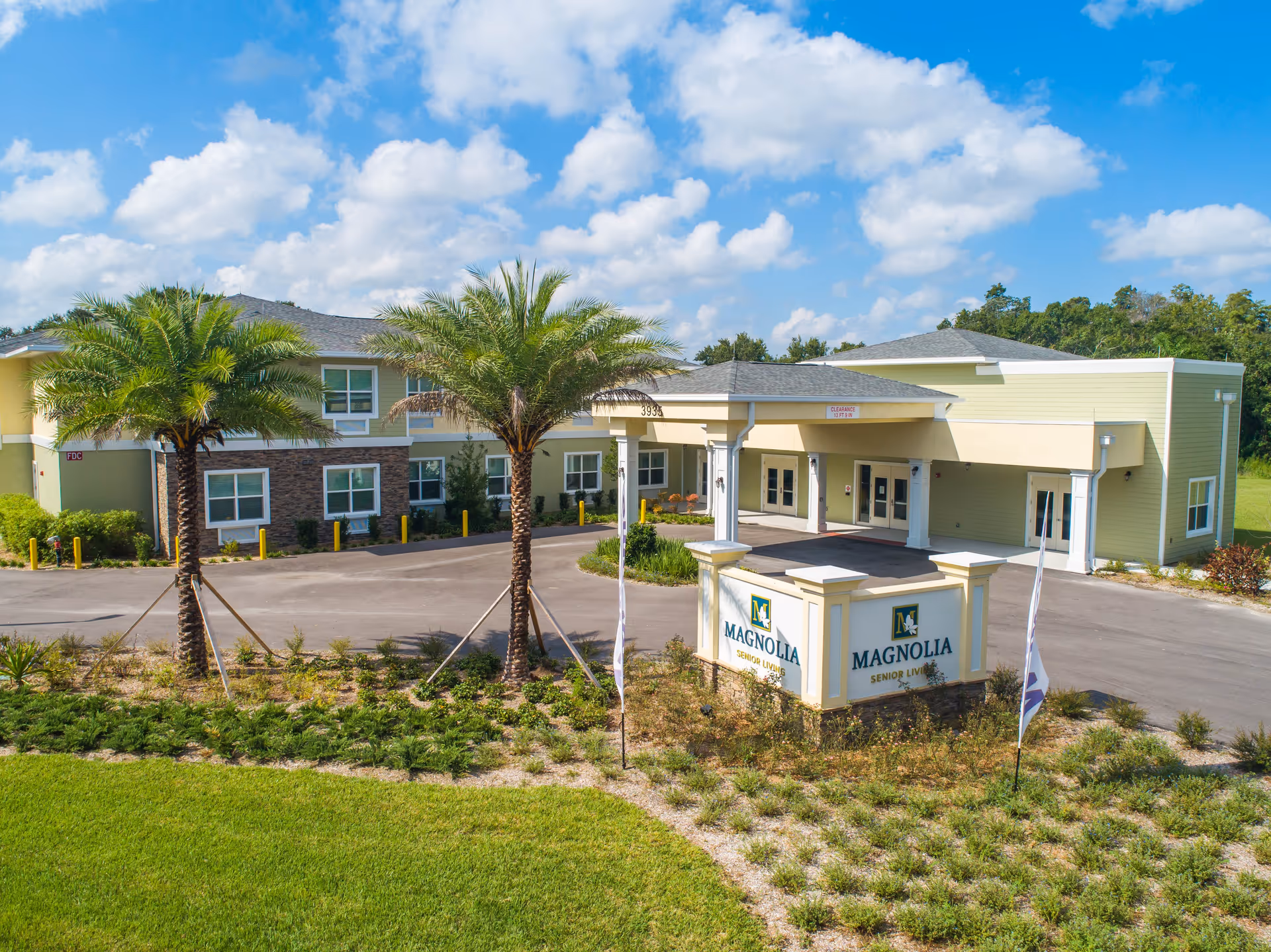 Exterior view of Magnolia Senior Living facility showing a two-story building with a covered entrance, palm trees, landscaped greenery, and a sign with the facility's name in front under a partly cloudy blue sky.