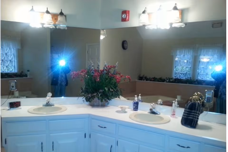 Double-sink bathroom vanity with a large mirror, floral centerpiece, toiletries, and overhead lights, with the photographer's flash reflected in the mirror.