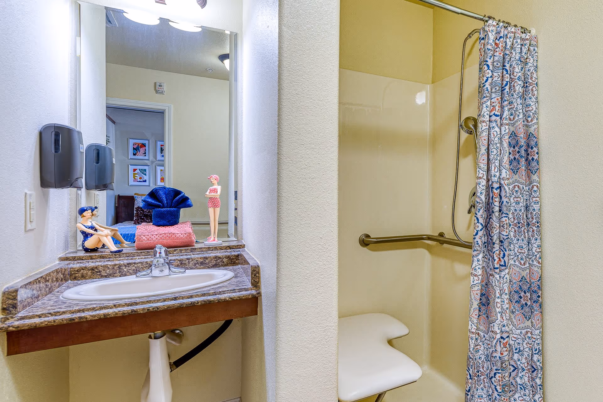 A bathroom area with a sink and countertop on the left, decorated with two small figurines and folded towels. On the right is a shower with a patterned curtain, a grab bar, and a built-in shower seat.