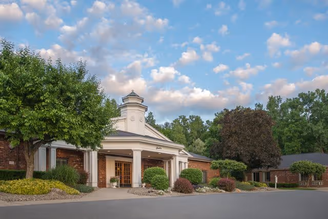 Exterior view of a senior living facility named Legacy at Grande Vie, featuring a brick building with a white entrance canopy, surrounded by well-maintained landscaping including trees and shrubs under a partly cloudy sky.