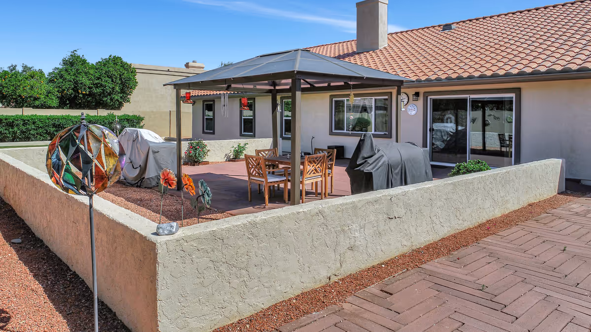 Outdoor patio area with a gazebo covering a wooden table and chairs, two covered grills, decorative garden ornaments, and a beige stucco wall surrounding the space. The building has a tiled roof and sliding glass doors leading inside.