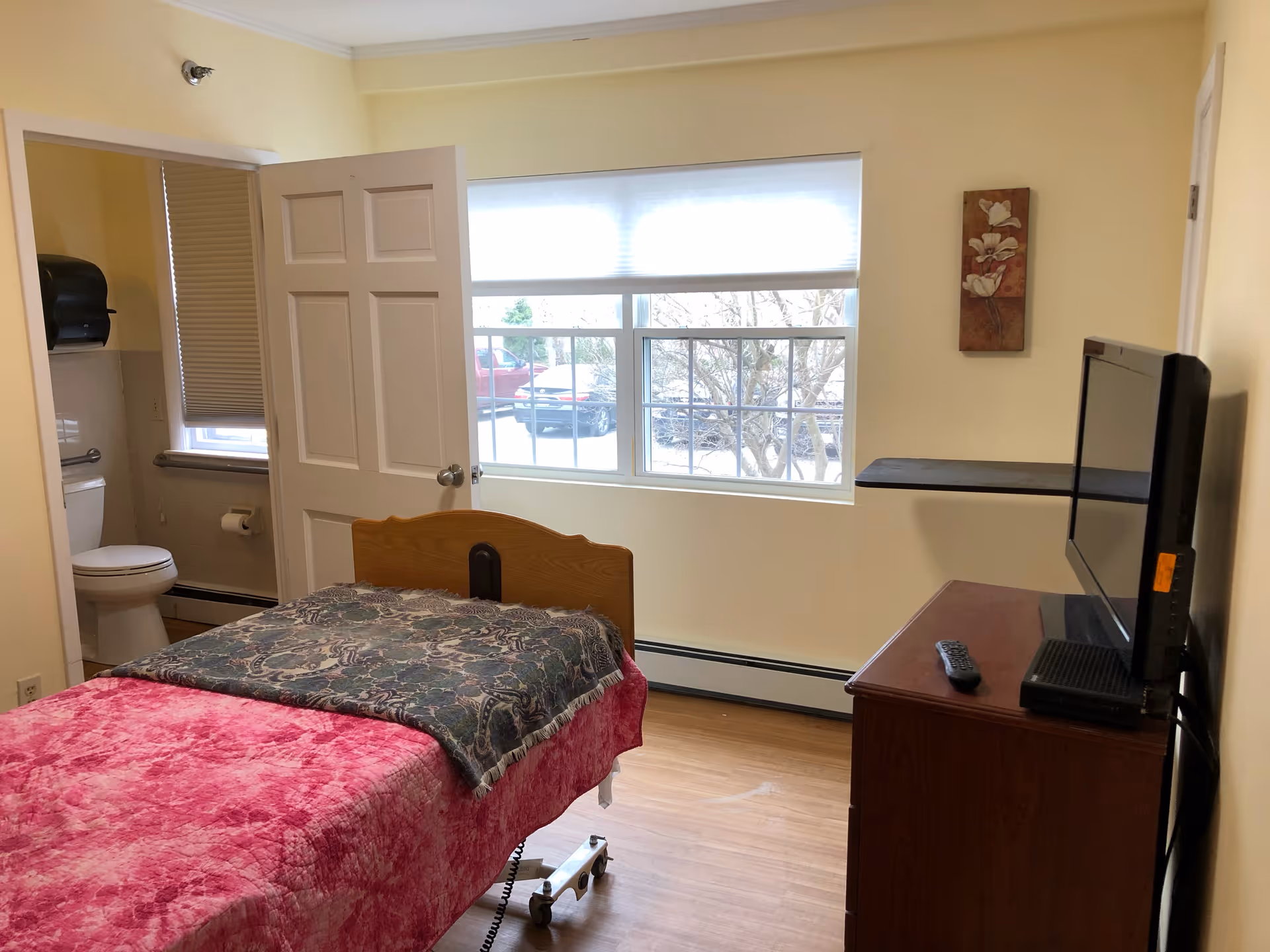 A small bedroom in a skilled nursing facility with a single bed covered in a pink blanket and a patterned throw. The room has a window with a view of parked cars and trees outside. There is a wooden dresser with a flat-screen TV and remote on top. The door to an adjacent bathroom is open, showing a toilet and a window with a closed blind.