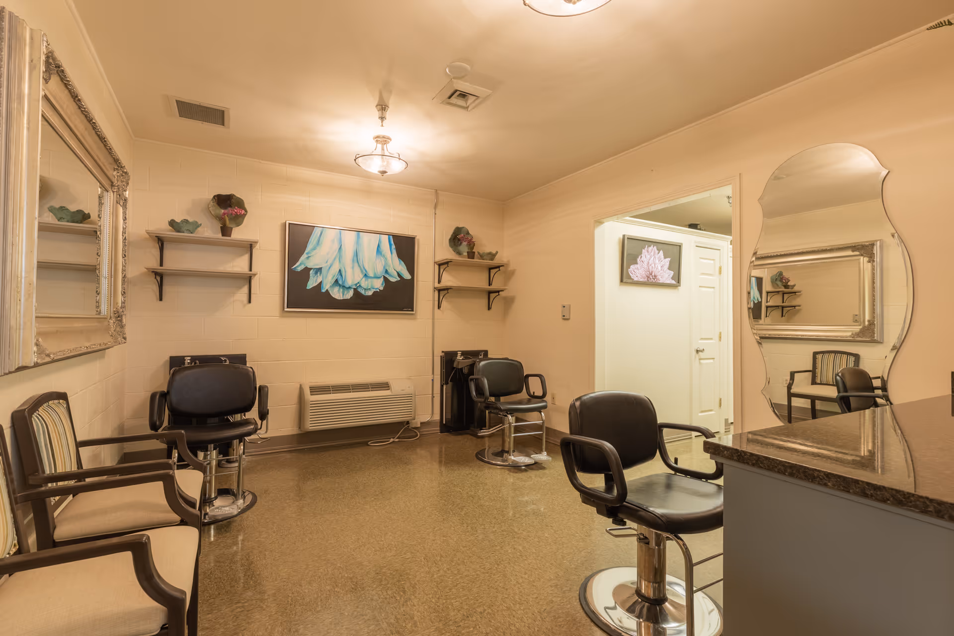 Interior view of a salon area in a senior living facility with three black salon chairs, two beige armchairs, a large ornate mirror on the left wall, a uniquely shaped mirror on the right wall, and a painting of a blue flower on the back wall. The room has beige walls, two ceiling lights, and shelves with decorative items.