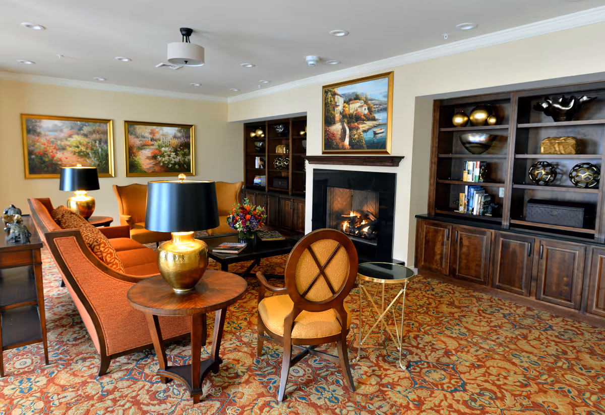 Cozy assisted-living common room with sofas, armchairs, decorative lamps, a fireplace, built-in bookshelves and framed paintings.