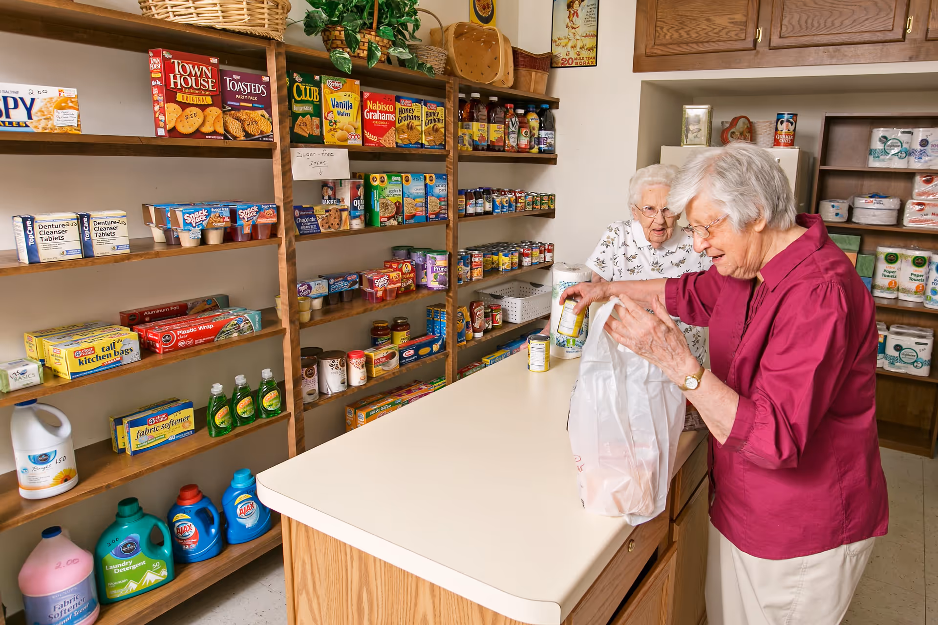 Two elderly women in a small store or pantry area with shelves stocked with various groceries and household items. One woman in a magenta shirt is placing canned goods into a plastic bag on a counter, while the other woman in a white floral blouse watches. The shelves behind them hold snacks, canned foods, cleaning supplies, and paper products.