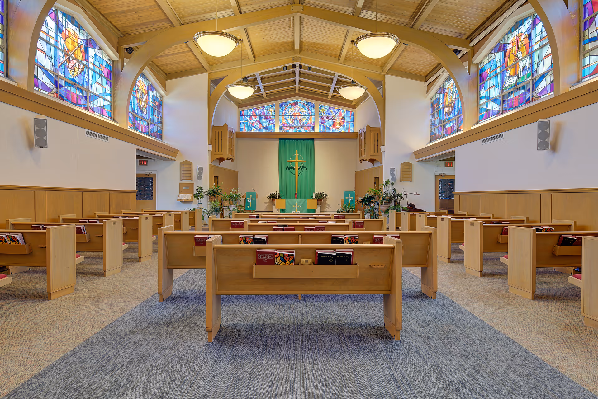 Interior view of a chapel with wooden pews arranged in rows facing an altar. The chapel features stained glass windows along the upper walls and a wooden vaulted ceiling with hanging light fixtures. The altar area has green cloth decorations and plants on either side.