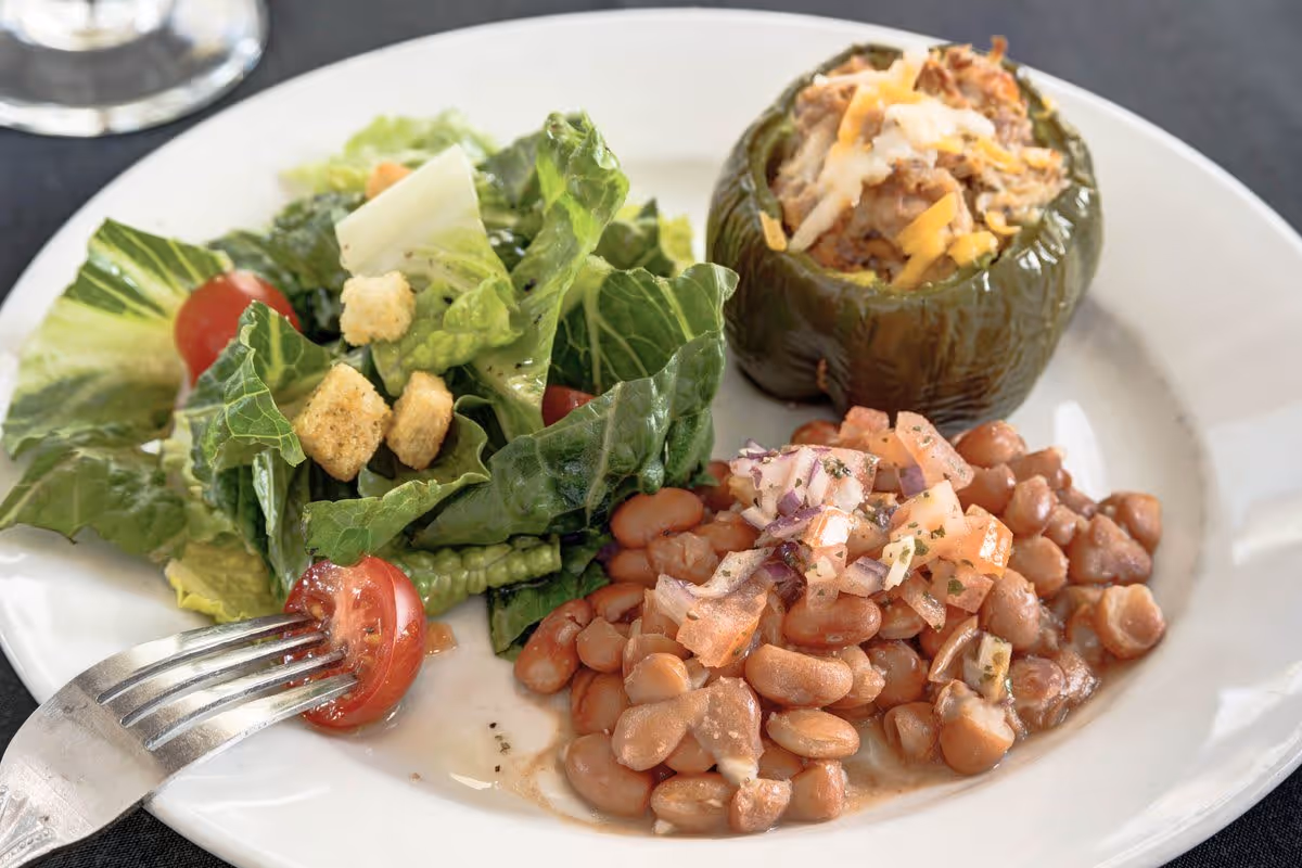 A white plate with a fresh green salad containing romaine lettuce, cherry tomatoes, and croutons, a serving of pinto beans topped with diced onions and tomatoes, and a stuffed green bell pepper filled with a cheesy meat mixture. A fork is placed on the plate near the salad.