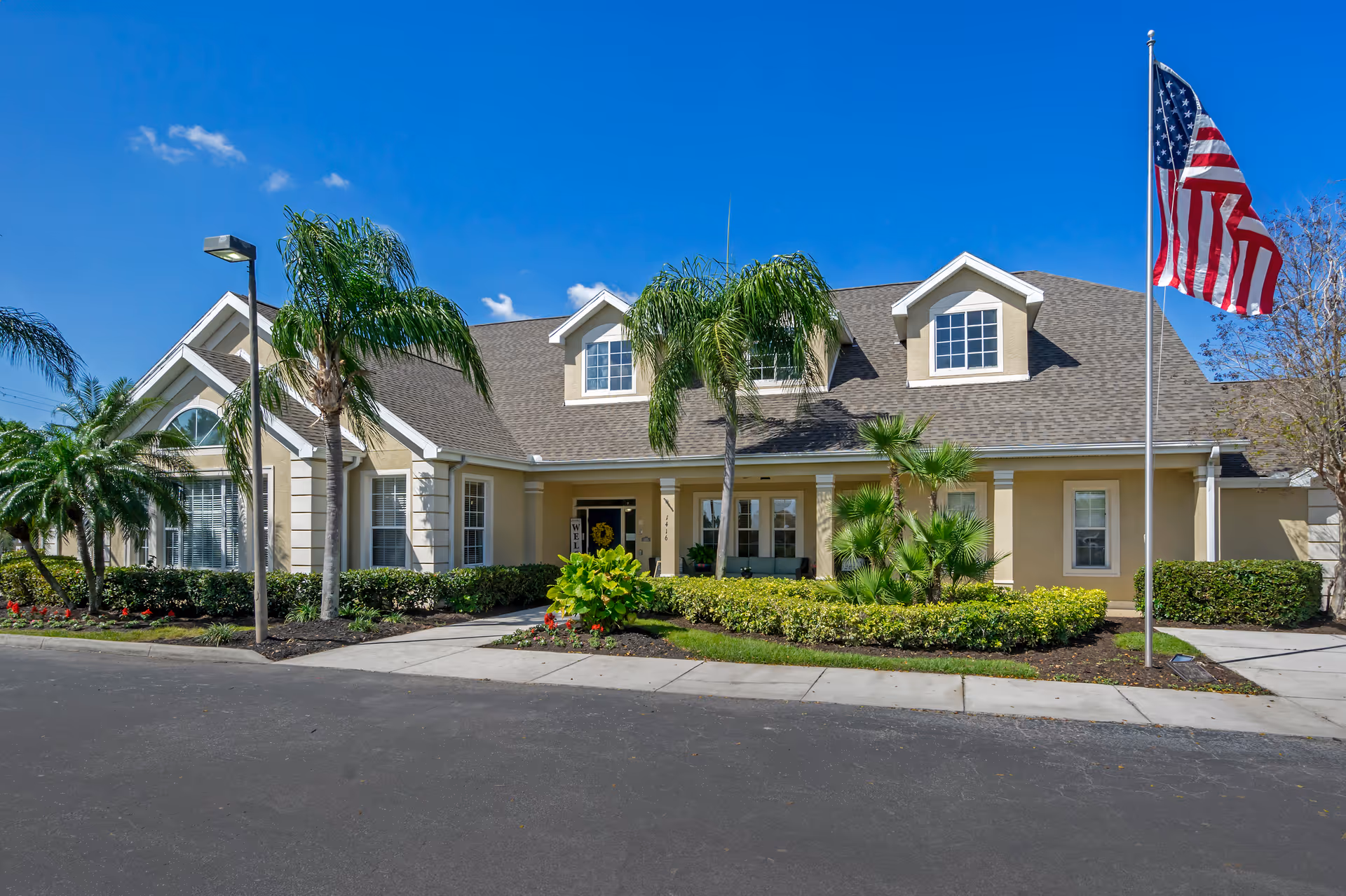 Exterior view of a single-story building with beige walls and a gray shingled roof under a clear blue sky. The building has multiple windows, a covered entrance with columns, and is surrounded by palm trees, shrubs, and landscaped flower beds. An American flag is flying on a flagpole to the right of the entrance.
