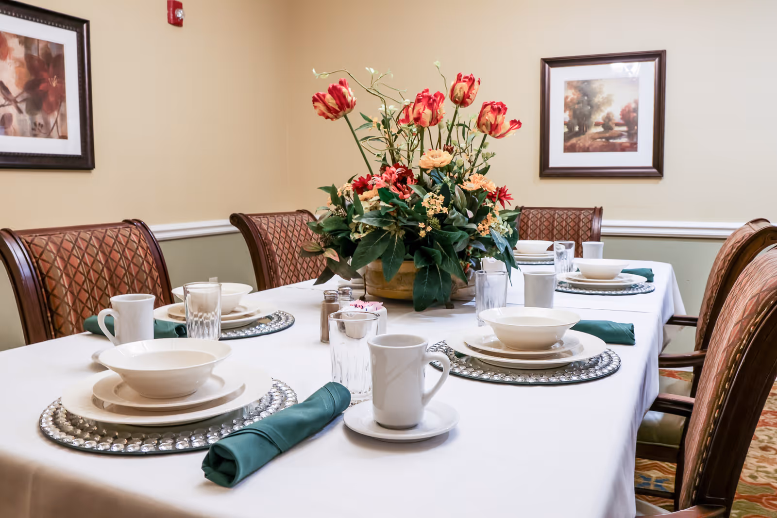 A dining table set for six with white tablecloth, white plates, bowls, cups, glasses, and green cloth napkins. A large floral centerpiece with red and orange flowers is in the middle of the table. The room has beige walls with two framed paintings and cushioned wooden chairs around the table.