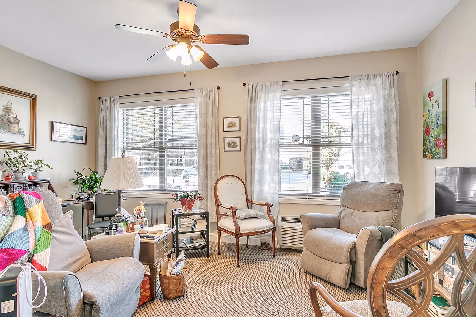 A cozy living room with two large windows covered by white curtains and blinds. The room features a ceiling fan with lights, two armchairs, a wooden chair with white upholstery, a small side table with a lamp, plants, framed artwork on the walls, and a TV on the right side.