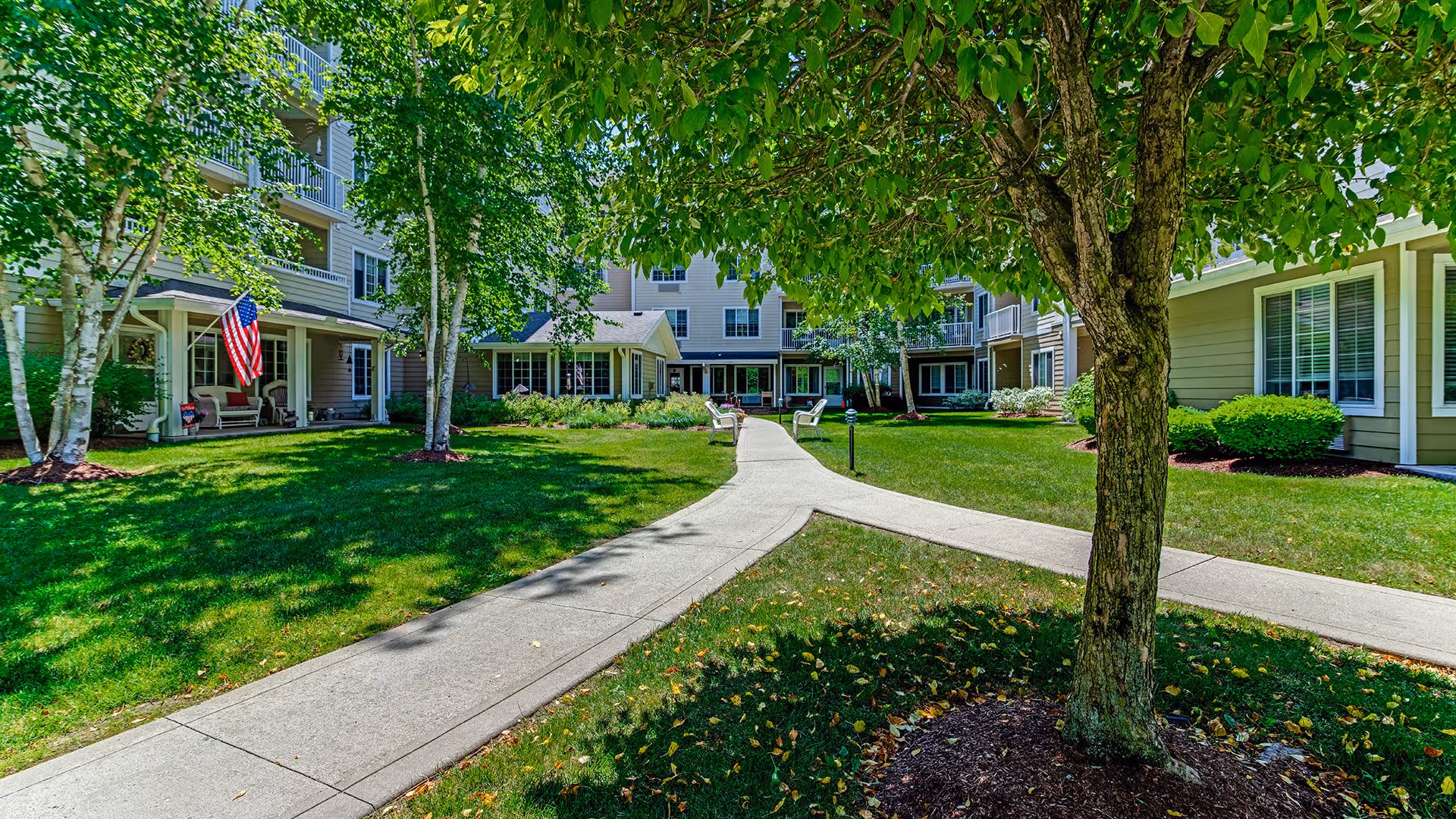 A sunny courtyard area at Holiday Birch Heights featuring a concrete walkway that splits into two paths surrounded by green grass and trees. The multi-story building with beige siding and white trim has balconies and windows overlooking the courtyard. There are benches along the walkway and an American flag displayed near the entrance of the building.