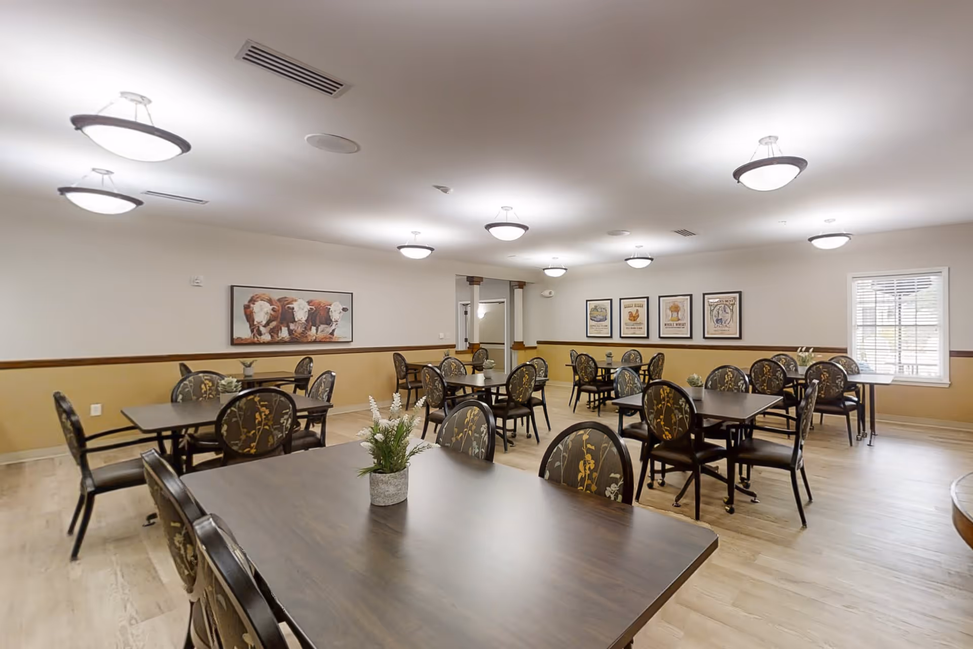 A spacious dining room with multiple dark wood tables and chairs featuring floral upholstery. The room has light-colored walls with a wooden trim and several framed pictures on the walls, including one of cows. Ceiling lights provide bright illumination, and a window with blinds allows natural light in.