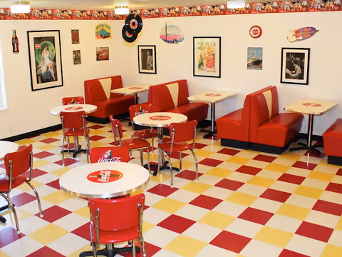 A retro-style dining room with red and yellow checkered floor tiles, red chairs, and red booth seating. The tables have Coca-Cola logos on them. The walls are decorated with vintage posters, signs, and records, creating a nostalgic diner atmosphere.