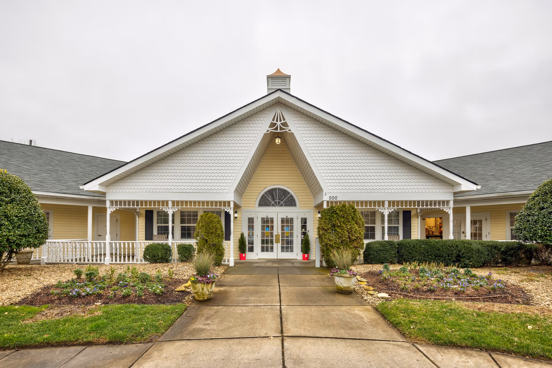Front exterior view of TerraBella Cramer Mountain facility with a paved walkway leading to double glass doors under a peaked roof with decorative white trim. The building is yellow with white accents and surrounded by landscaped bushes, flower beds, and grass.