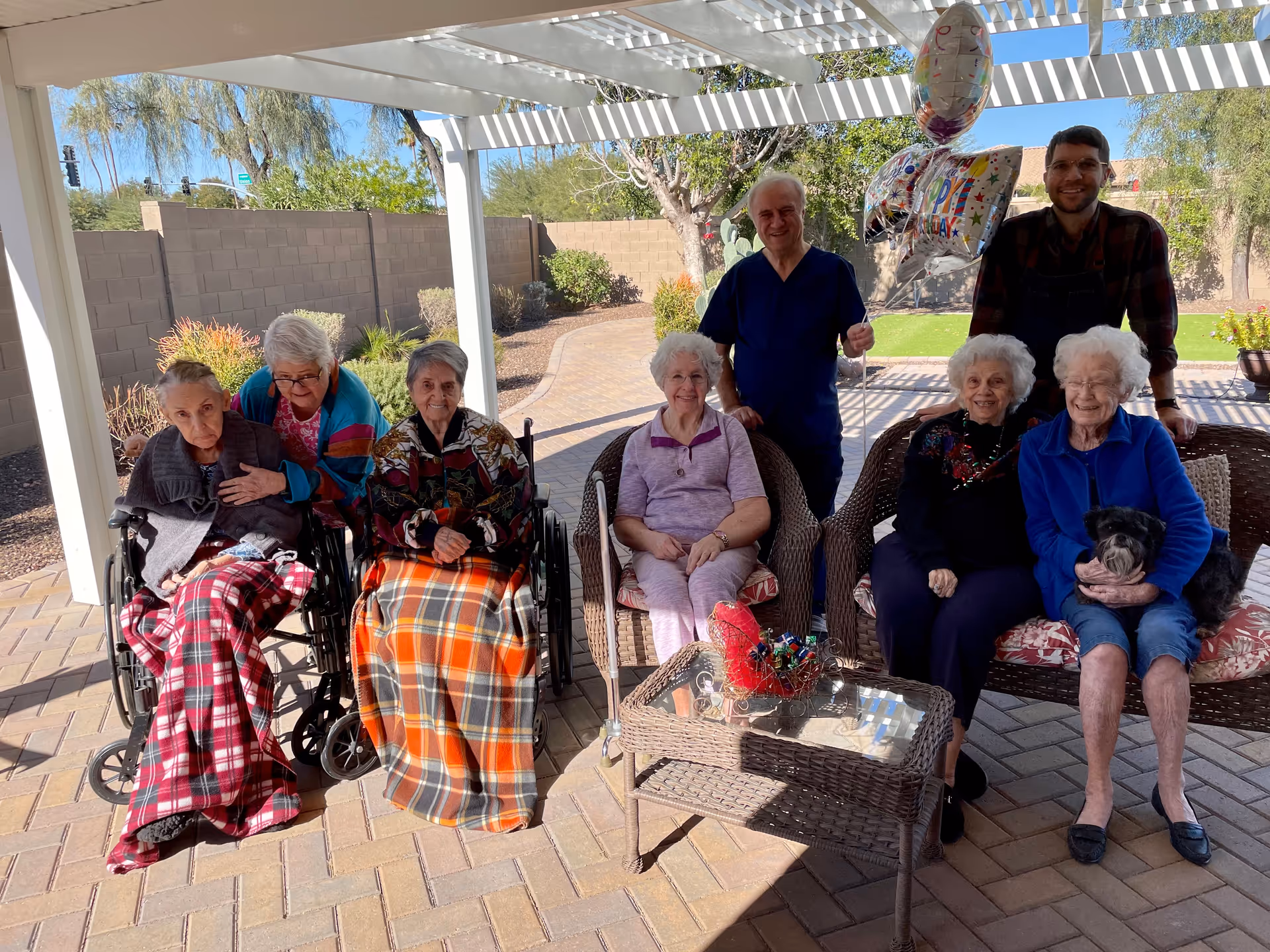 A group of elderly women and two men are gathered outdoors under a white pergola. Four elderly women are seated on wicker chairs and two in wheelchairs, some covered with blankets. One elderly woman is holding a small dog. One man is standing behind the group holding birthday balloons, and another man stands beside him. The setting is a paved patio with plants and a brick wall in the background.