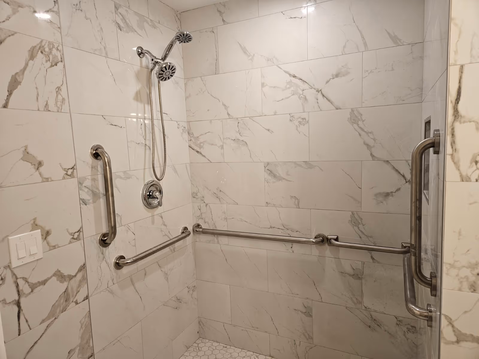 Accessible shower stall with marble-look tile walls, a handheld showerhead, and stainless steel grab bars.