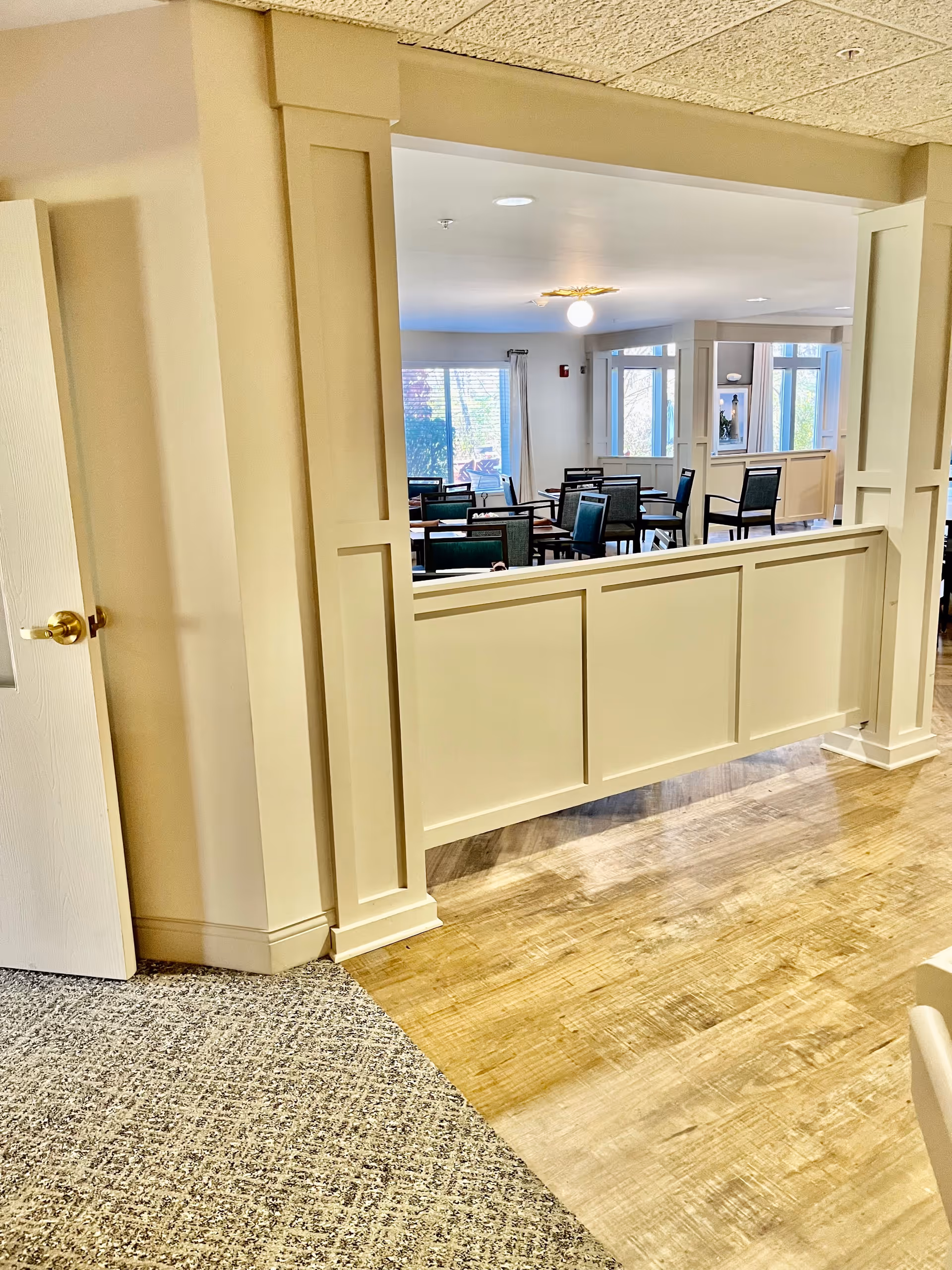 Interior view of a senior living facility showing a dining area with multiple tables and chairs. The room has large windows letting in natural light, beige walls, and a mix of carpet and wood flooring. A partial wall with decorative paneling separates the hallway from the dining space.