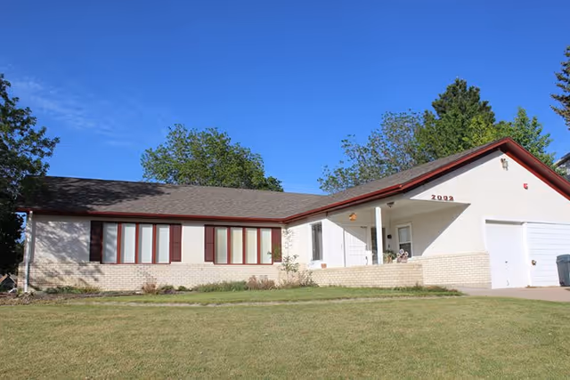 Single-story residential building with a light-colored exterior, red trim, and a sloped roof under a clear blue sky. The building has multiple windows with red shutters and a garage on the right side. There is a grassy lawn in front and some trees in the background.