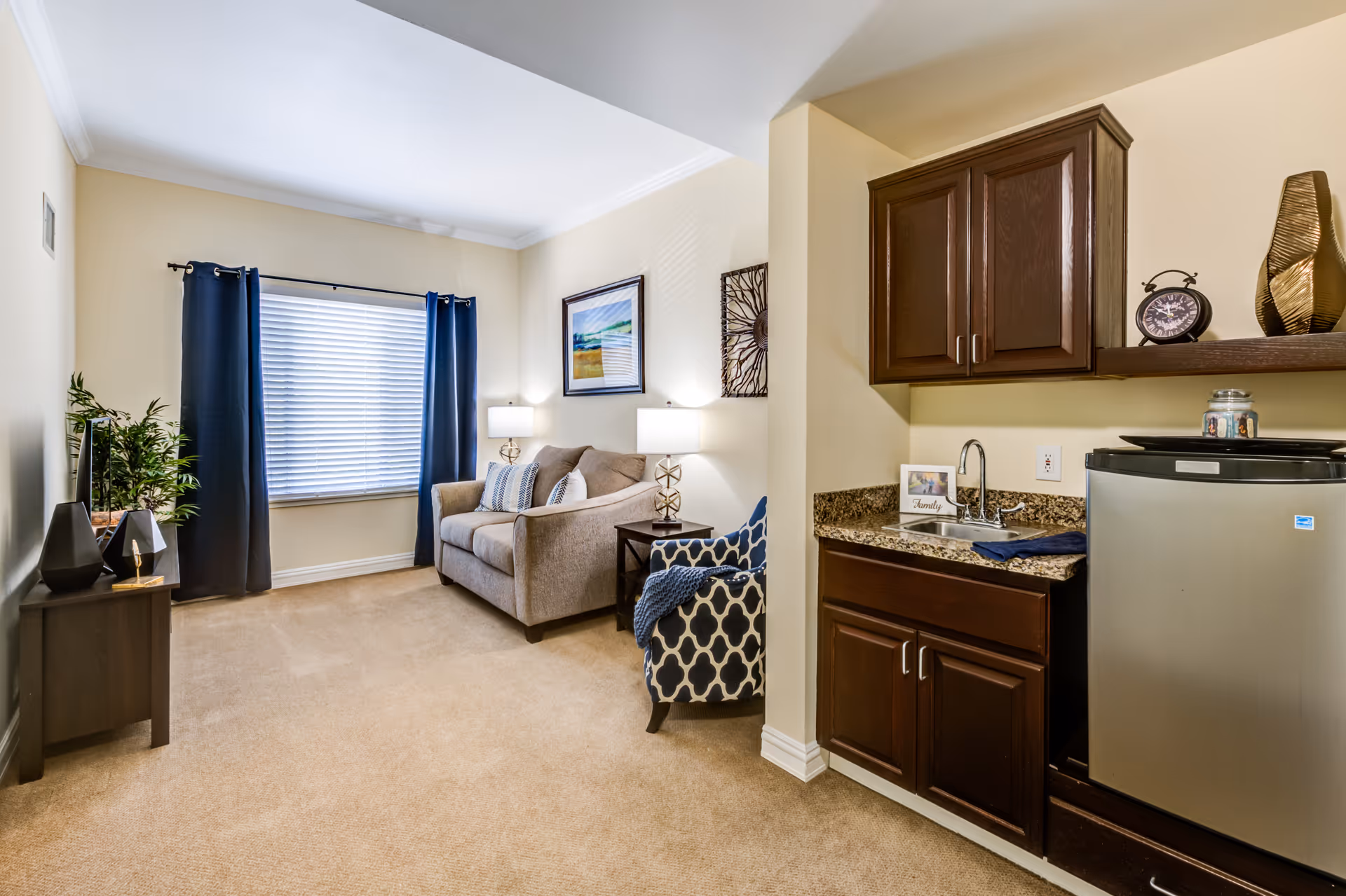 A cozy senior living room with beige carpet and walls, featuring a beige loveseat with patterned pillows, a blue and white patterned armchair, two table lamps on either side of the loveseat, and a window with blue curtains and white blinds. To the right, there is a small kitchenette area with dark wood cabinets, a granite countertop, a small sink, and a stainless steel mini fridge. Decorative items including a clock and a vase are placed on the shelf above the kitchenette.