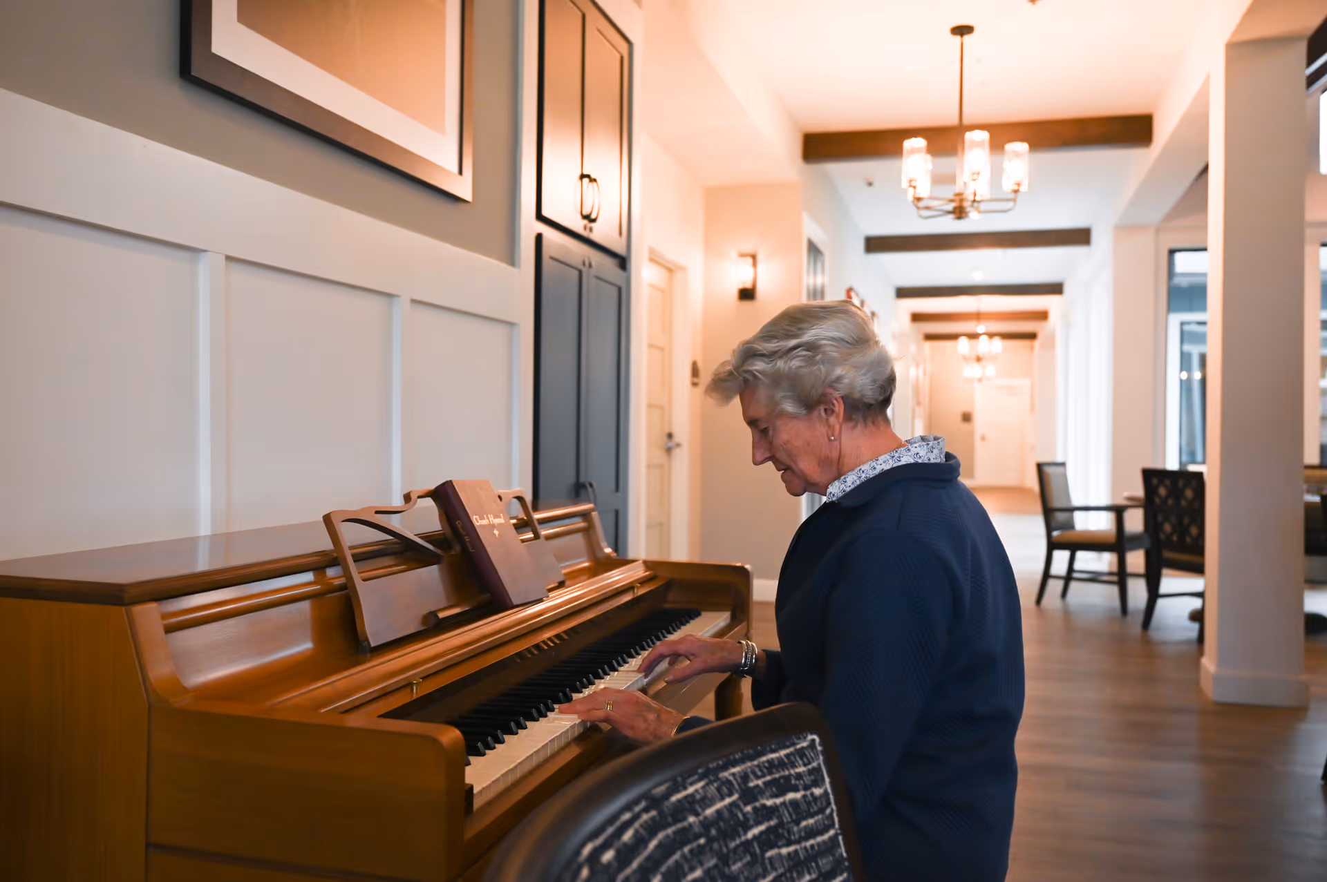 An elderly woman playing a wooden piano in a well-lit hallway with modern decor, including chandeliers, chairs, and framed artwork on the walls.