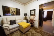 A cozy living room with a cream-colored leather sofa adorned with a dark pillow and a yellow throw blanket on an ottoman in front. The room features a patterned area rug, a table lamp on a side table, a decorative vase with dried branches, and framed artwork on the walls. An open doorway leads to a hallway with additional furniture visible.