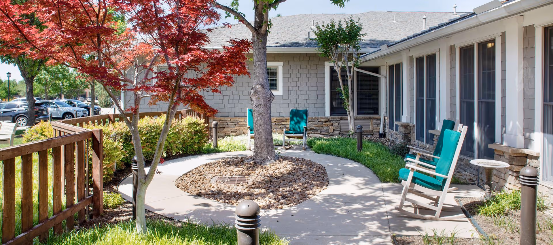 Outdoor courtyard area at Sunrise of Fort Worth featuring a circular concrete pathway around a tree with rocks at its base, surrounded by green grass and bushes. There are several wooden rocking chairs with teal cushions placed along the pathway near the building with large windows. A wooden fence and parked cars are visible in the background.