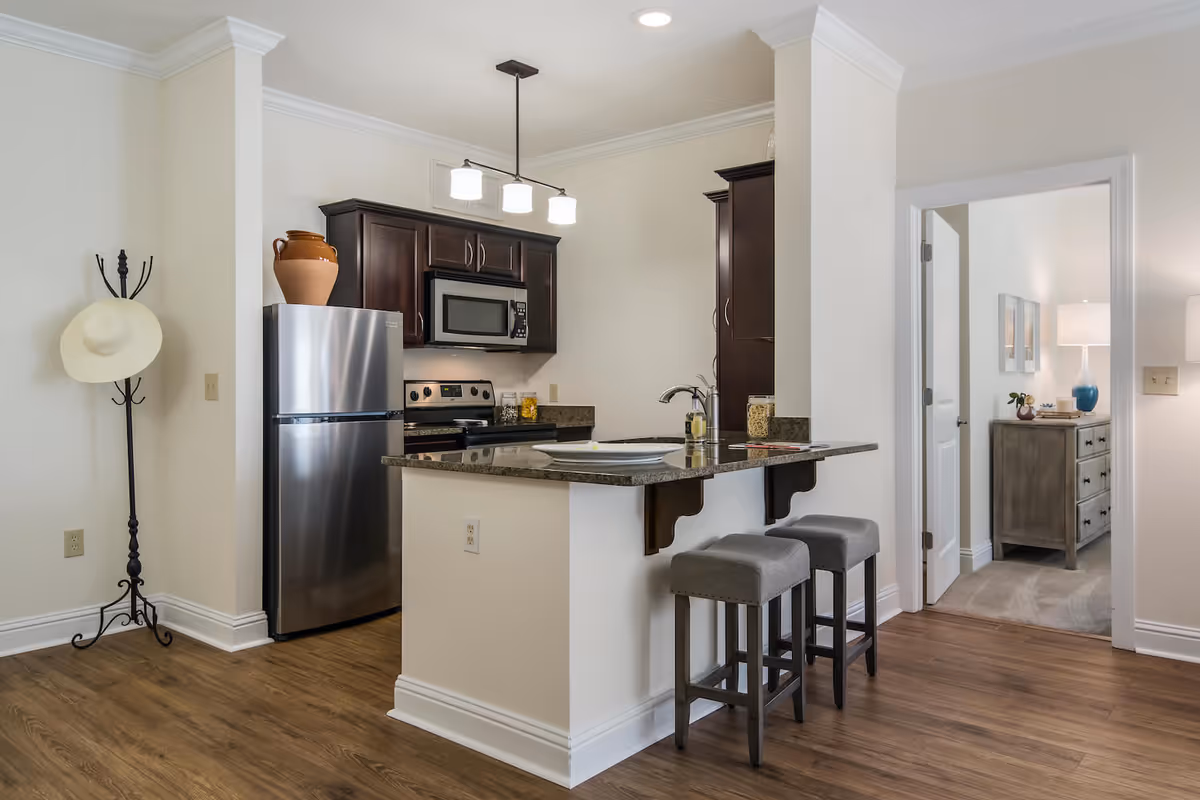 Modern kitchen area with dark wood cabinets, stainless steel refrigerator, microwave, and stove. A kitchen island with granite countertop has two gray cushioned bar stools. A coat rack with a hat is placed against the wall, and an open doorway leads to a room with a wooden dresser and a lamp.