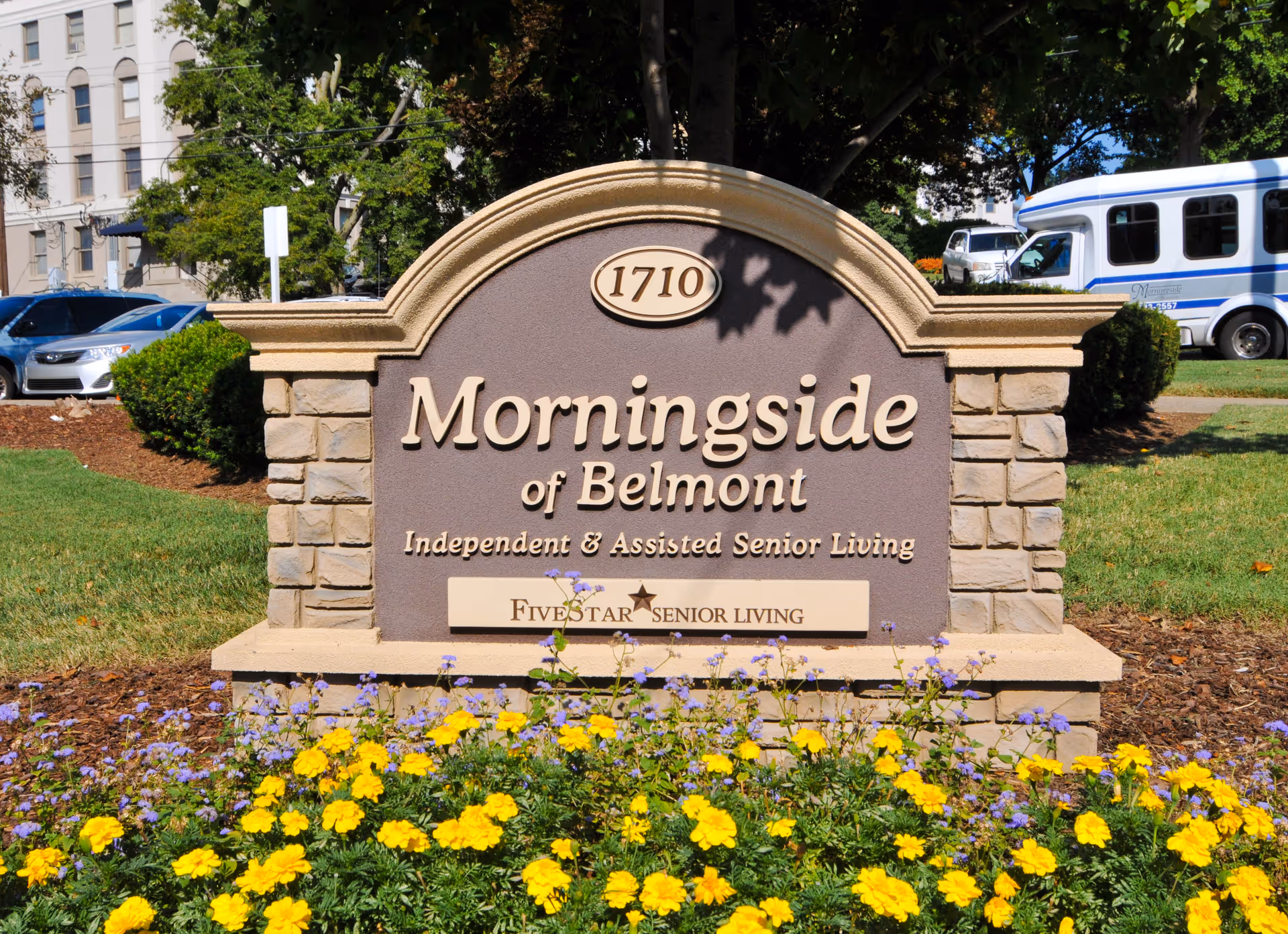 Stone and concrete sign for Morningside of Belmont, an independent and assisted senior living facility, surrounded by yellow and purple flowers with parked cars and a shuttle bus in the background.