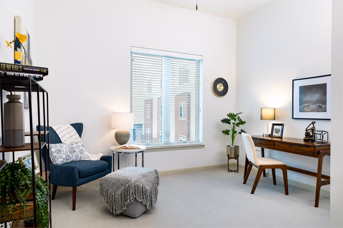 A bright and cozy room with a blue armchair adorned with a floral pillow and a striped throw blanket. Next to the chair is a small round side table with a lamp and an open book. Across from the chair is a wooden desk with a white cushioned chair, a table lamp, framed pictures, and decorative items. A large window with blinds lets in natural light, and a green potted plant is placed near the desk. The walls are white, and the carpet is light gray.