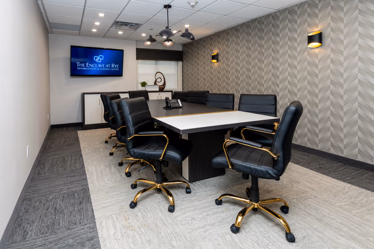 A modern conference room with a rectangular table surrounded by black leather office chairs with gold accents. The room has a patterned gray wall with two wall sconces, a ceiling light fixture, and a mounted flat-screen TV displaying 'The Enclave at Rye Rehabilitation & Nursing Center'. There is a window with a blind and a decorative sculpture on a cabinet beneath the TV.