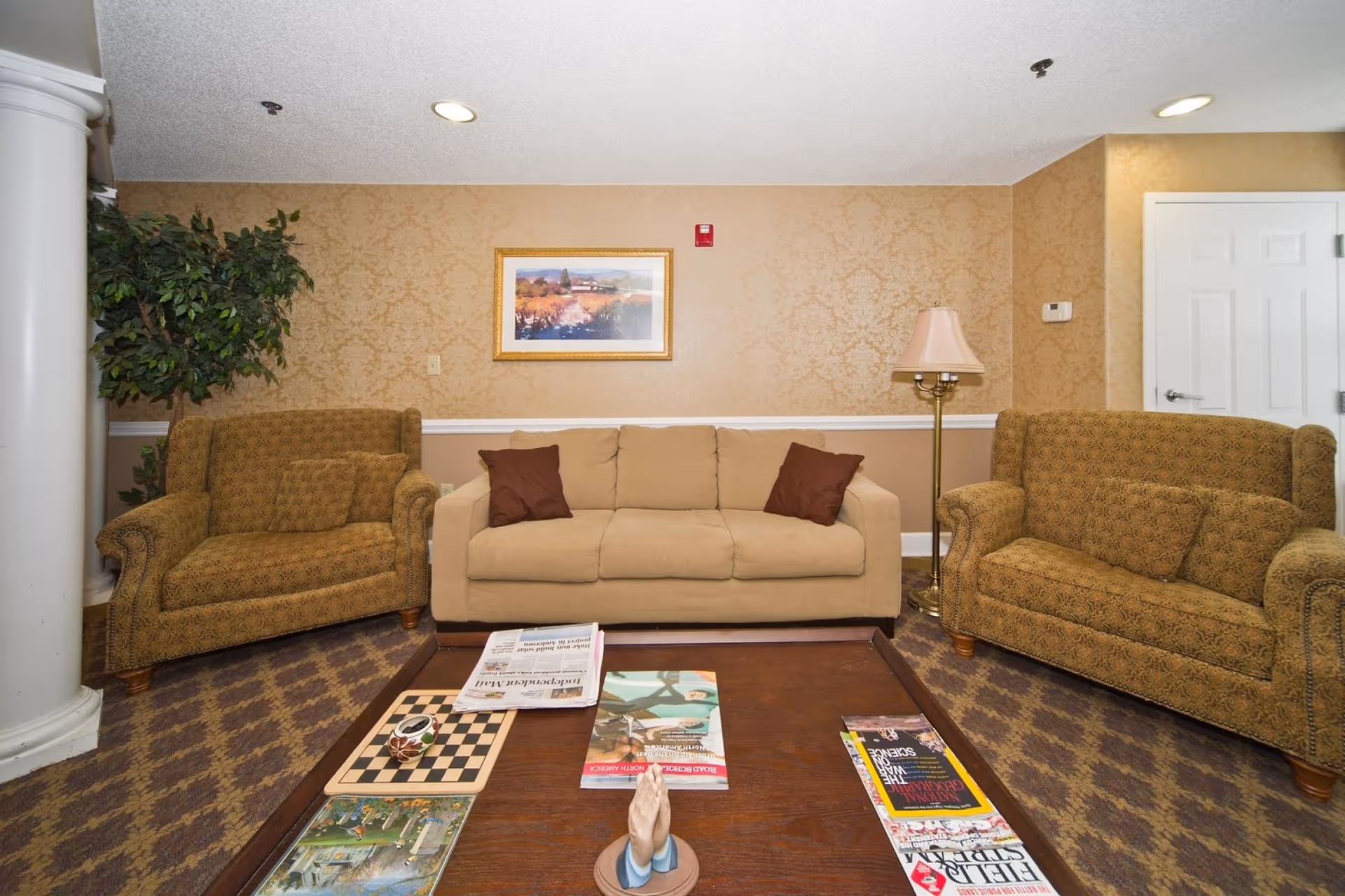 A cozy living room area with a beige sofa flanked by two patterned armchairs. A wooden coffee table in front holds magazines, a chessboard, and a small decorative item. The walls have a textured beige wallpaper with a framed landscape painting hanging above the sofa. A floor lamp stands to the right, and a large potted plant is on the left near a white column.